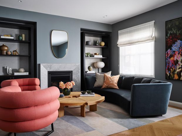 A stylish living room with dark charcoal built-in shelving and cabinetry, a marble fireplace surround, grey-blue walls, and a large window with a white Roman blind. The room features a curved coral-red lounge chair, a dark blue sofa and armchair, a round timber coffee table, decorative cushions, a geometric area rug, and a colourful floral artwork. The overall scheme is polished, contemporary, and richly layered with curved furniture forms.