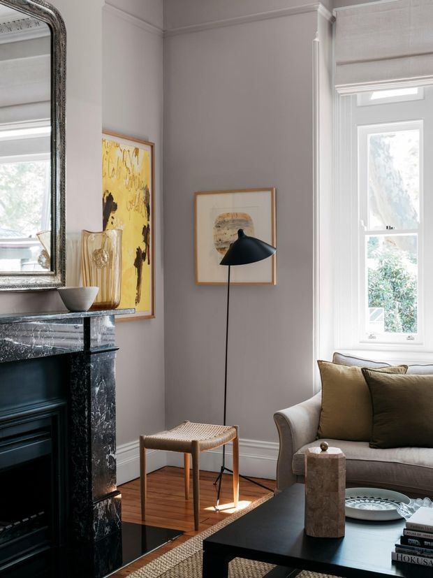 A refined sitting room with pale grey walls, decorative ceiling and wall mouldings, a black marble fireplace with ornate mirror, tall white-trimmed window, contemporary black floor lamp, framed artwork, a cushioned armchair, and a dark coffee table. Warm timber flooring and restrained neutral furnishings create a sophisticated traditional-meets-contemporary interior.