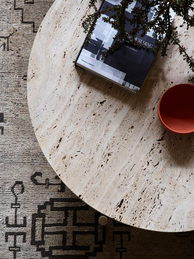 Overhead close-up of a round cream travertine coffee table with pronounced porous veining, placed over a patterned black-and-beige rug. A dark art or architecture book and a small terracotta-red bowl sit on the tabletop, with leafy branches entering from the top edge.