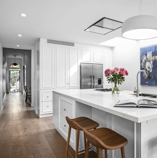 Bright, refined kitchen with white raised-panel cabinetry, a large white island with integrated seating, stainless-steel appliances, pale timber flooring, and a contemporary pendant light. A hallway extends beyond the kitchen, and a framed artwork adds colour to the otherwise neutral space.