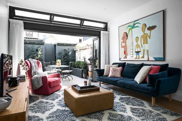 A bright, furnished living room with a deep blue sofa, red accent armchair, patterned area rug, square upholstered coffee table, timber media console, and large framed artwork. Full-height glazed doors and clerestory-style windows open toward a compact courtyard, while white curtains and neutral walls keep the room airy.
