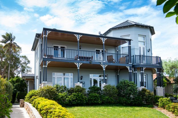 Exterior view of a two-storey light-coloured home with a prominent covered upper and lower verandah, dark decorative railings and posts, traditional sash-style windows, a hipped roof, and landscaped front garden.