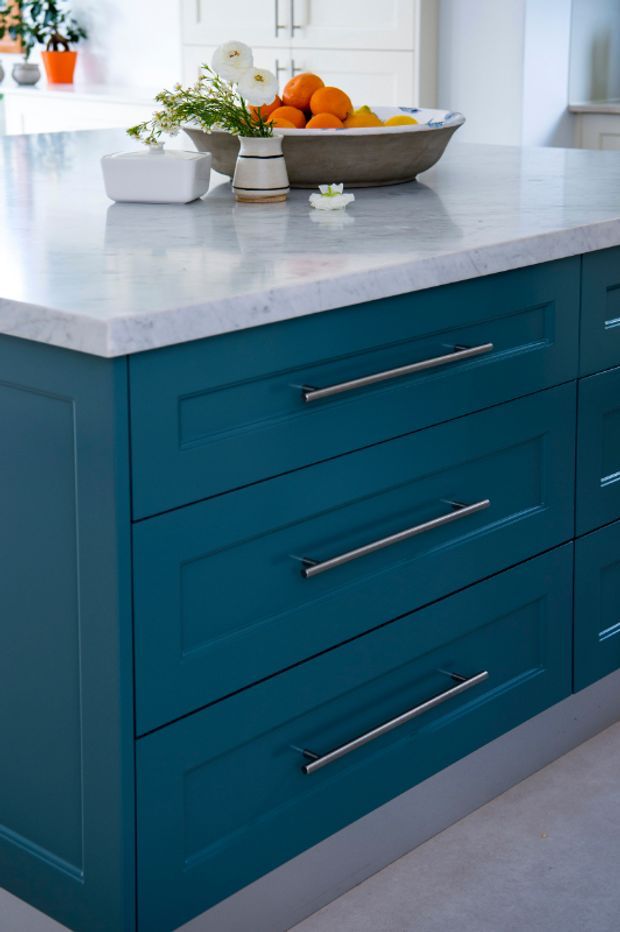 Close-up of a contemporary kitchen island with deep teal-blue shaker-style cabinetry, long brushed metal handles, and a pale veined stone benchtop. A large decorative bowl of fruit and a small floral arrangement sit on the island, with white kitchen cabinetry visible in the background.