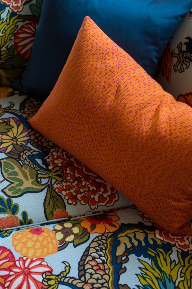 Close-up interior detail of a richly patterned upholstered seating surface with a vivid orange textured throw pillow in the foreground and a deep teal-blue cushion behind it. The surrounding upholstery features dense multicoloured floral and botanical motifs, creating a bold, eclectic decorative composition.