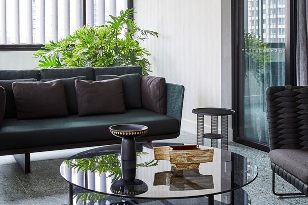 A sophisticated contemporary living room in a high-rise urban setting, featuring a dark teal upholstered sofa with charcoal cushions, sculptural black accent tables, a reflective glass coffee table, and a large leafy indoor plant. Floor-to-ceiling glazing and vertical windows create a bright, refined atmosphere with city buildings visible beyond.