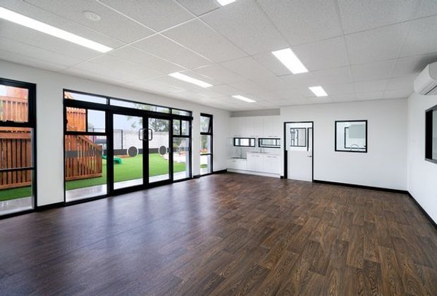 A bright, spacious contemporary commercial or multipurpose interior with dark wood-look flooring, white walls, a suspended acoustic ceiling with recessed linear lighting, extensive black-framed glazing and doors, and simple white built-in cabinetry. The glazing looks onto a fenced landscaped outdoor area. An air-conditioning unit and wall-mounted screen are visible.