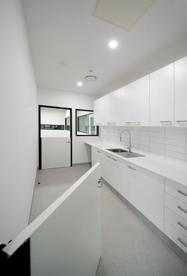 Bright, compact white kitchenette or utility kitchen with extensive flat-panel cabinetry, a white benchtop, stainless-steel sink and tap, and a white horizontal subway-tile splashback. The room has a pale grey resilient-looking floor, recessed ceiling lights, black-framed internal glazing and a partially open foreground door or partition.