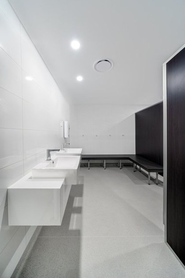 A clean, minimalist commercial washroom or changing-room interior with white tiled walls, floating white basins and integrated fixtures along the left wall, pale grey seamless flooring, recessed ceiling lights, and dark timber-look privacy partitions with bench seating along the right and rear walls.