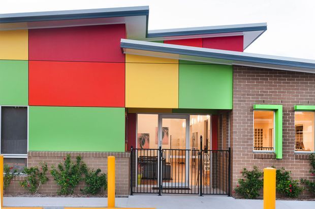 Exterior view of a colourful contemporary low-rise building with bold red, yellow and green geometric wall panels, exposed brickwork, projecting metal roof planes, bright green window surrounds, and a gated entrance. Landscaping and bollards are visible in front of the facade.
