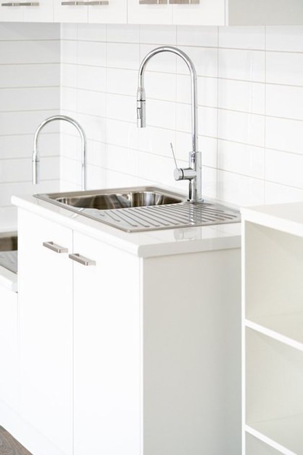 Bright, compact white kitchen featuring a stainless-steel sink with integrated draining board, tall curved chrome gooseneck tap, white tiled splashback, and white flat-panel cabinetry with slim metallic handles. Open white shelving is visible at right.