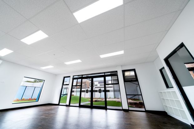Bright, empty commercial-style interior with white walls, a suspended acoustic ceiling and recessed fluorescent lighting. The room has dark wood-look flooring, extensive black-framed glazed doors and windows, and a white built-in open storage unit. Exterior landscaping and neighbouring buildings are visible through the glazing.
