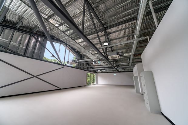 An expansive, mostly empty commercial interior with a polished light floor, white walls, and a highly visible exposed industrial ceiling. Black steel structural beams, exposed ductwork, cable trays, conduits, suspended circular lights, and rooftop mechanical services dominate the ceiling. Large windows and glazed panels bring in daylight, while angular black framing details appear along the left partition wall. Built-in white storage or service cabinets line the right wall.