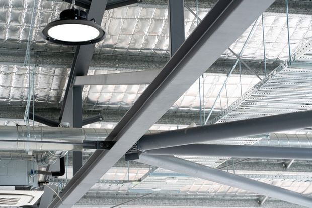 Close-up view of an exposed commercial ceiling and building-services zone, showing grey structural steel beams, suspended cable trays, ductwork, pipes, reflective foil insulation, and a round pendant light. The image emphasizes visible infrastructure and utilitarian construction detailing.