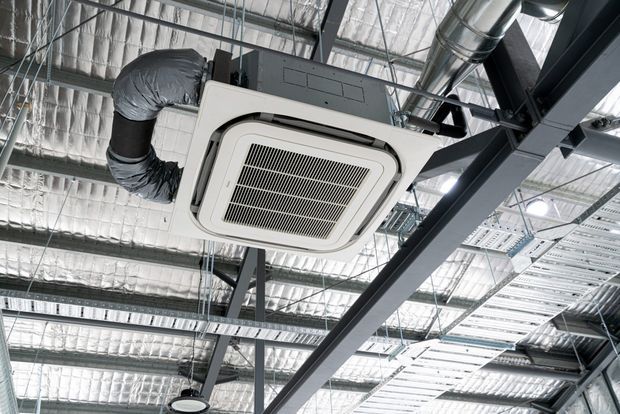Close-up view of an exposed commercial ceiling with a suspended cassette air-conditioning unit, silver ductwork, reflective insulation, cable trays, and dark steel structural framing.
