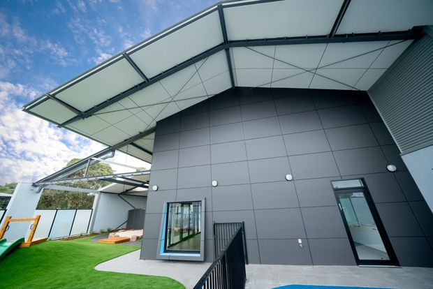 Low-angle exterior view of a contemporary building with a dramatic oversized curved steel-framed roof canopy, dark grey panel cladding, tall glazed openings, and a landscaped artificial-grass courtyard. A covered outdoor area and neighbouring building elements are visible to the side.