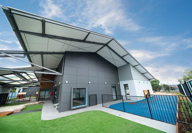 Exterior view of a contemporary low-rise building with a dramatic angular folded roof, dark grey panelled facade, extensive covered outdoor structures, and a fenced bright-blue pool or play-area feature. Artificial green lawn and paved paths surround the building.