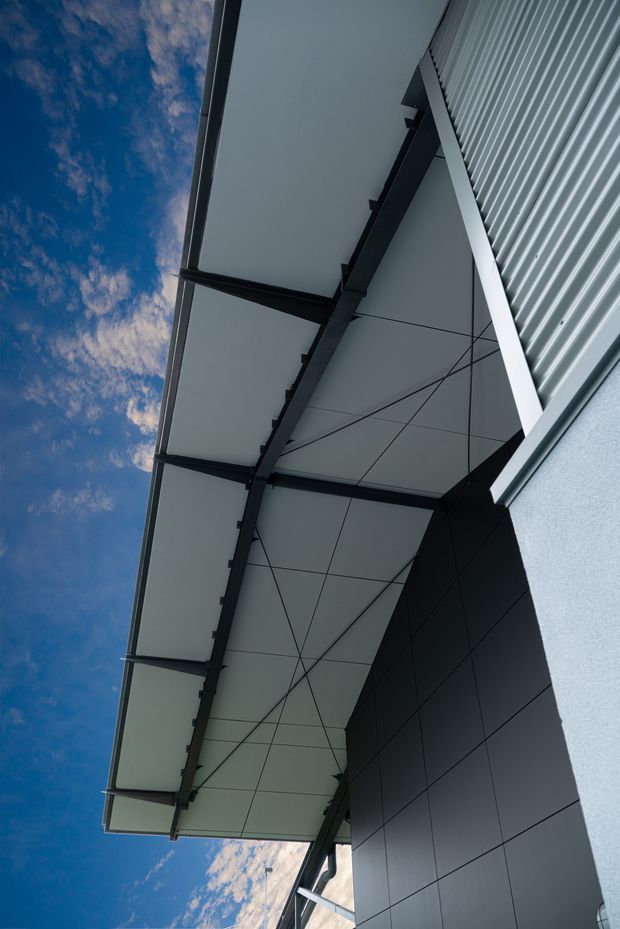 Upward-looking architectural detail of a contemporary building exterior, showing a broad projecting canopy with dark steel framing, smooth light soffit panels, dark rectangular wall cladding, and corrugated metal roofing. The composition emphasizes strong geometric lines and structural supports against a partly cloudy blue sky.