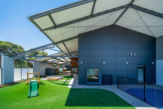 Exterior view of a contemporary low-rise building with a dramatic angular, extended roof canopy, dark blue panelled walls, and covered outdoor circulation. The landscaped grounds include bright artificial turf, a children's slide and play equipment, with black metal balustrades and glazed openings along the building.