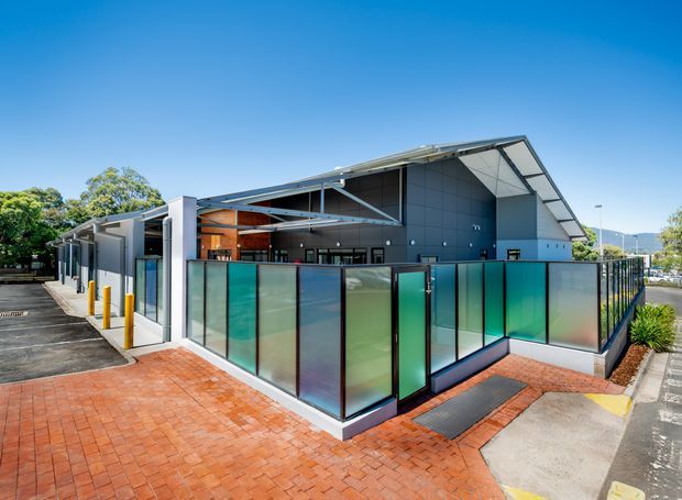 Exterior view of a contemporary low-rise commercial building with a prominent angular pitched roof, dark metal cladding, exposed steel framing, extensive glazing, and translucent teal privacy screens. The building is set beside a paved pedestrian area with landscaped edges and accessible entry paving.