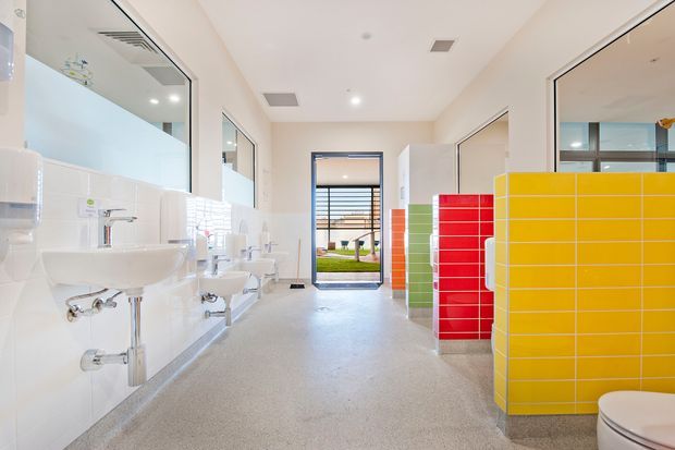 Bright contemporary communal bathroom or washroom with multiple wall-mounted white basins, exposed chrome plumbing, white tiled walls, a light grey seamless floor, and vivid tiled privacy partitions in yellow, red, orange, and green. Full-height glazing and a glazed door bring in daylight, with an outdoor lawn area visible beyond. The space has a clean, functional institutional or commercial fit-out.