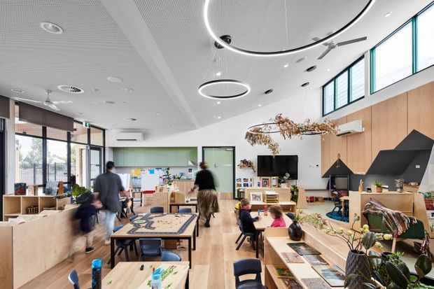 Bright contemporary communal interior, likely a childcare or educational activity room, with low timber storage and work tables, children and adults, a television and shelving, large glazed windows, timber wall panelling, and distinctive circular suspended ceiling lights. The space is open, airy, and carefully fitted out for shared learning and play.