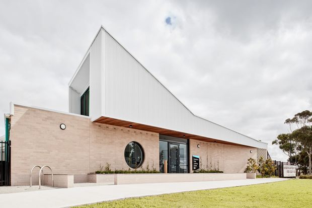 Exterior view of a contemporary low-rise building with a dramatic steep triangular roof form, white vertically ribbed metal cladding, pale brick masonry walls, dark-framed glazing, timber-lined soffit, and a circular window. The building fronts a broad paved entry area and lawn under an overcast sky.