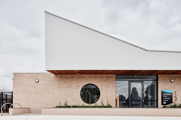Contemporary single-storey building facade with pale brick walls, a distinctive sloping white corrugated roof form, warm timber-lined soffit, circular window, and dark glazed entrance doors. A sign identifies the building as a learning centre. The image is photographed from outside under an overcast sky.