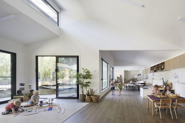 Bright, open-plan educational or childcare interior with a large play area in the foreground, dining and activity tables in the background, extensive glazing, high white ceilings, and a long built-in kitchenette or storage wall. The space has warm timber-look flooring, black-framed windows and doors, and abundant natural light.