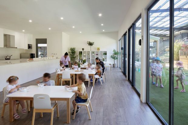 Bright contemporary childcare dining and activity room with multiple low timber tables and white child-sized chairs, children and caregivers seated for meals, a minimalist white kitchen with island at left, open shelving and potted plants, and extensive dark-framed glazed doors opening to an outdoor play area. Light timber-look flooring and recessed ceiling lights create an airy, clean interior.