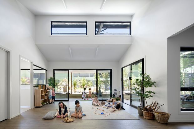 Bright, spacious double-height family room with extensive black-framed glazing, clerestory windows, pale timber flooring, white walls and a large rug. Children are playing in the open space, with built-in or freestanding timber shelving and indoor plants visible around the perimeter. The room opens toward a landscaped outdoor area through large glass doors.
