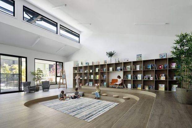 Bright, expansive double-height reading and activity space with full-height glazing, clerestory windows, built-in timber shelving filled with books and objects, and a curved built-in seating area surrounding a large rug. Several children are seated on the floor, while lounge chairs and indoor plants add softness to the open, contemporary interior.