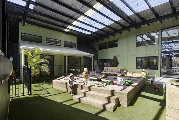 A spacious indoor-outdoor children's play area within a large glazed, steel-framed structure. The space features a translucent pitched roof, pale green wall panels, high-level windows, artificial turf flooring, and several raised timber sandpit or planter-like play structures with steps. Children are using the central sand and play area, while a covered veranda and planting are visible along the rear.