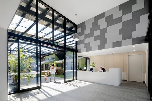 Bright contemporary commercial reception or lobby with a dramatic double-height glazed entrance and internal courtyard-like atrium. Black-framed floor-to-ceiling glazing, grey tiled flooring, a large white reception desk, pale timber wall joinery, pendant lighting, and a geometric grey-and-white acoustic feature wall define the space. The open glazed roof structure brings in abundant daylight and greenery is visible beyond the entrance.