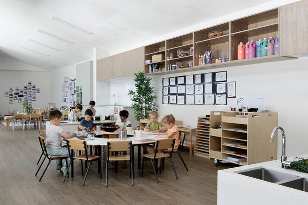Bright, spacious educational classroom or childcare studio with children seated around shared tables. The room has warm wood-look flooring, white walls and ceiling, extensive open wood storage cabinetry, shelves of colourful supplies, display boards, a large stainless-steel sink, and abundant natural light. The overall atmosphere is clean, practical, creative, and welcoming.