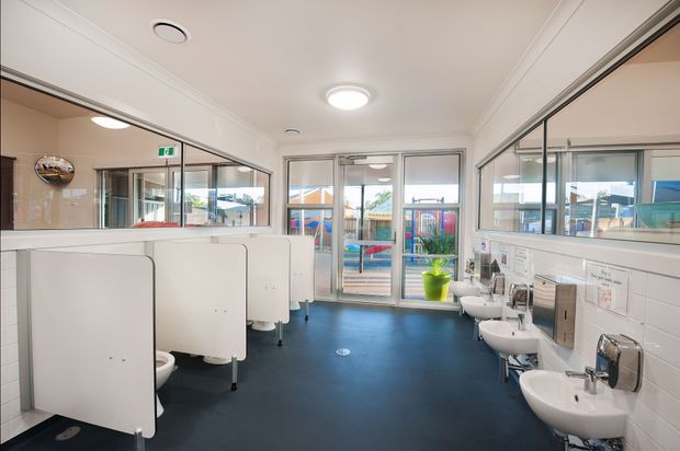 Bright, clean commercial bathroom with multiple white toilet cubicles on the left and wall-mounted basins along the right. The room has dark blue resilient flooring, white tiled walls, large mirrors, fluorescent-style ceiling lighting, and glazed doors/windows opening toward an outdoor play area.