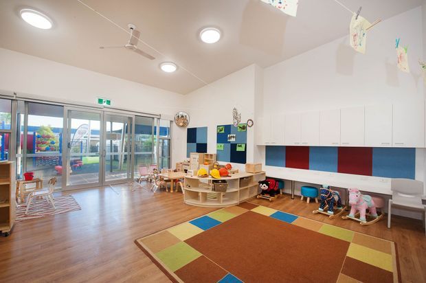 A bright, spacious childcare or preschool activity room with polished timber-look flooring, colourful geometric area rug, low child-sized tables and chairs, toy storage, wall-mounted cabinetry, and playful artwork. Full-height glazed sliding doors open to an outdoor play area. The room has a high sloping ceiling with ceiling fans and recessed lighting.
