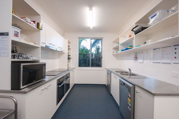 A bright, narrow galley-style kitchen with white cabinetry, open shelving, stainless-steel appliances, grey benchtops, a white tiled splashback, and a dark blue resilient floor. A central window brings in natural light, while the space also includes a microwave, oven, dishwasher, sink, and practical storage.