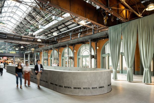 A large adaptive-reuse industrial interior, likely a public market or commercial hall, with a curved raw concrete reception or service counter in the foreground. The space features exposed steel trusses, ductwork, suspended industrial lighting, timber-toned structural elements, tall arched windows, and full-height sage-grey curtains. People and retail activity are visible in the background.