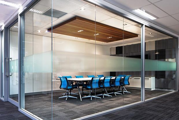 Contemporary commercial meeting room enclosed by full-height glass partitions with frosted privacy bands. The room contains a long white conference table with blue office chairs, dark carpet flooring, a compact kitchenette or storage joinery at the rear, and a striking timber slatted ceiling feature within a suspended ceiling grid.