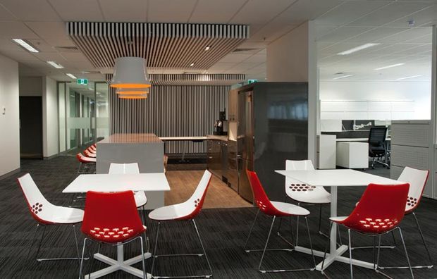 A contemporary commercial office breakout and collaboration area with several white tables and distinctive red-and-white chairs. The space includes a dark carpeted floor, timber-toned flooring near a kitchenette or service counter, glass partitions, open-plan workstations, and decorative timber slatted ceiling and wall features. Pendant lighting and recessed office lighting provide a bright, functional atmosphere.