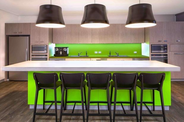 A contemporary kitchen with a large white island and five black bar stools. The kitchen features extensive light wood cabinetry, integrated stainless-steel appliances, a vivid lime-green splashback and matching island front, dark timber-look flooring, and three oversized dark pendant lights.