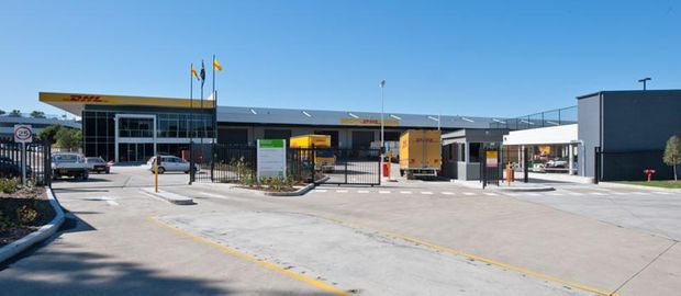 Wide exterior view of a contemporary commercial or light-industrial facility, with a low flat-roofed building, dark grey and yellow facade elements, security fencing and gates, concrete vehicle forecourt, loading or service bays, and clear blue sky. Flags and signage are visible near the entrance.