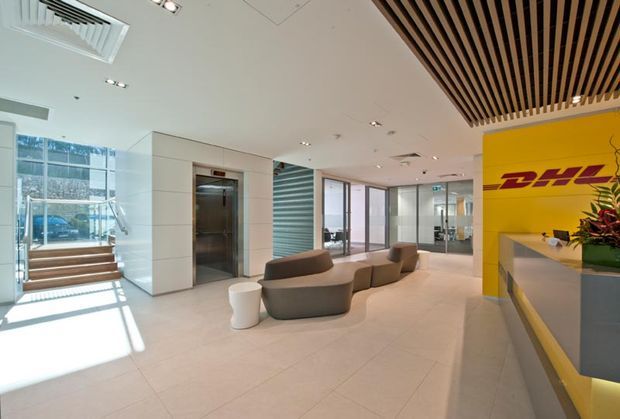 Bright, contemporary commercial reception lobby with a DHL-branded yellow feature wall and reception desk, sculptural upholstered seating, a lift, large glazed entry areas, pale tiled flooring, and a timber slatted ceiling feature.