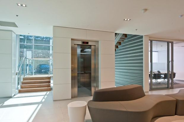 Bright contemporary commercial lobby or reception area with a central stainless-steel elevator, white tiled or panelled walls, polished light flooring, sculptural brown upholstered seating, and a glazed meeting room. A stair and large windows bring in natural light, while a ribbed green feature wall adds texture.