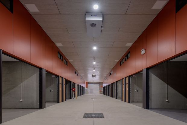 Long, symmetrical interior corridor in a modern commercial or institutional building, lined on both sides with enclosed rooms or bays behind dark-framed glazed and solid doors. The space has a broad pale concrete floor, exposed grey concrete wall sections, extensive rust-red overhead wall cladding or cabinetry, and a suspended acoustic tile ceiling with recessed lights and mechanical services. The corridor terminates at a roller or sectional door, suggesting a service, storage, workshop, or parking-related facility.