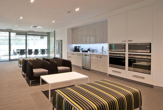 Open-plan contemporary living room with an integrated kitchen at the rear. The space features white built-in cabinetry, a vertically textured timber-toned kitchen backsplash, integrated stainless-steel appliances, recessed ceiling lighting, dark upholstered armchairs, and a large striped ottoman in black, grey, and yellow. Glazed doors connect the living area to a secondary workspace or dining area.