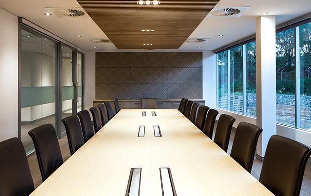 Contemporary corporate boardroom with a long light-wood conference table, integrated cable or power outlets, dark leather chairs, glazed partitions, expansive windows, a patterned feature wall, and a slatted timber ceiling panel with recessed lighting.
