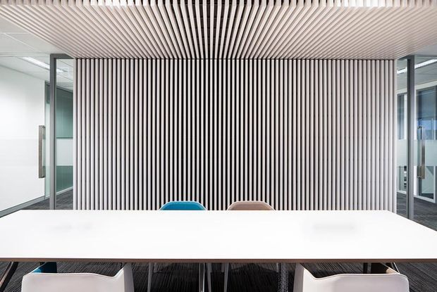 A bright contemporary commercial meeting room with a long white conference table and partially visible chairs. The focal feature is a full-height wall and ceiling treatment of closely spaced vertical timber-look slats, creating a ribbed linear pattern. Glazed partitions and glass doors flank the space, with carpeted flooring and a clean, minimalist office fit-out.