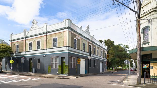 Street-level view of a two-storey heritage commercial corner building with an ornate parapet, decorative finials, rendered masonry facade and tall sash-style windows. The lower level is painted dark grey with a bright green entrance door, while the upper level has pale cream and muted red detailing. Adjacent urban buildings, utility poles, overhead wires and marked streets frame the scene.
