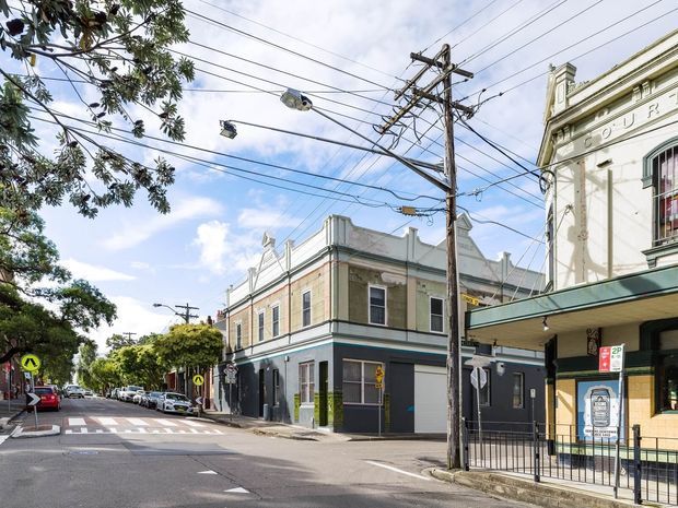Street-level view of a heritage-style urban corner building with ornate parapets, moulded facade details, tall sash-like windows, and a dark painted lower level. A neighbouring commercial frontage, overhead utility wires, pedestrian crossing, parked cars, and mature street trees line the streetscape.